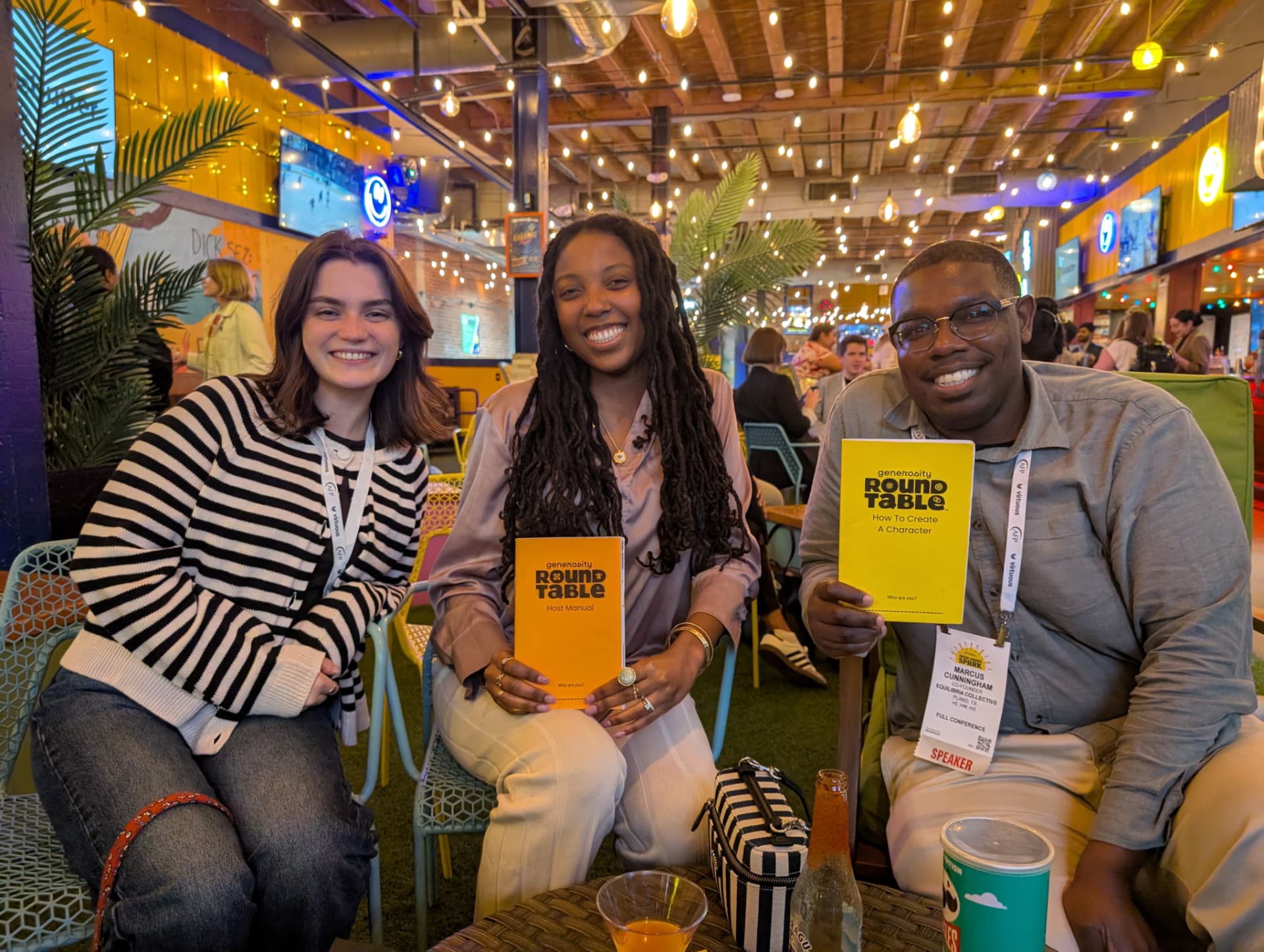 Three players smile while holding the Host Manual and How To Create A Character booklets at a bar.