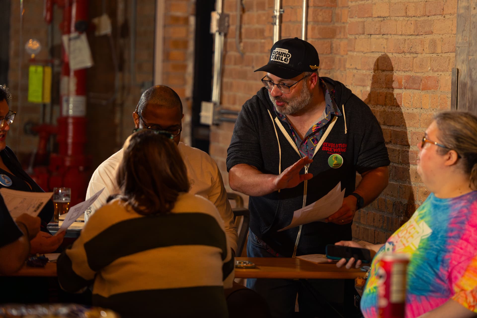 A Host in a Roundtable cap gestures while guiding players seated at a brewery table.