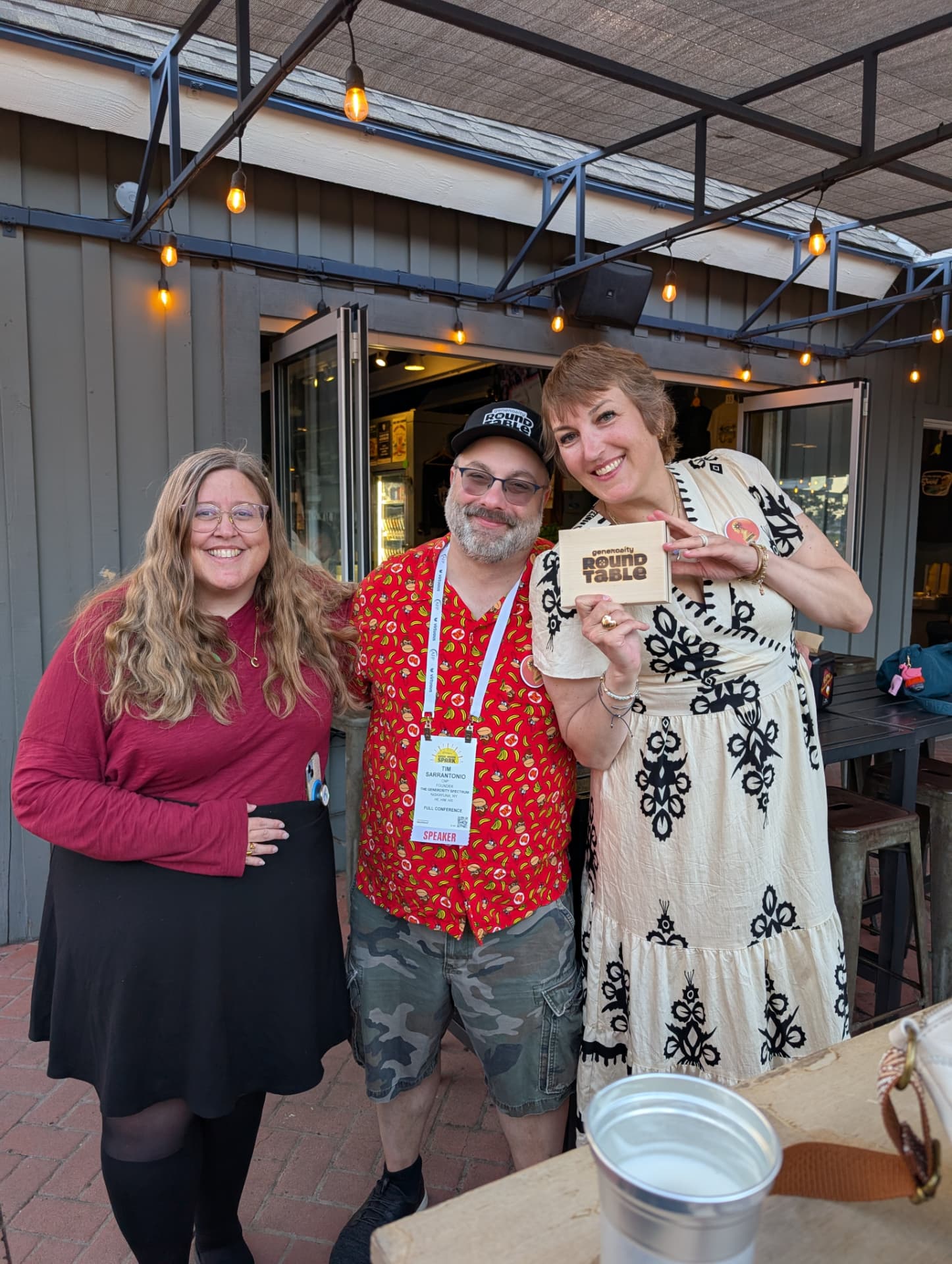 Three people smile together outdoors, one holding the wooden Generosity Roundtable box.