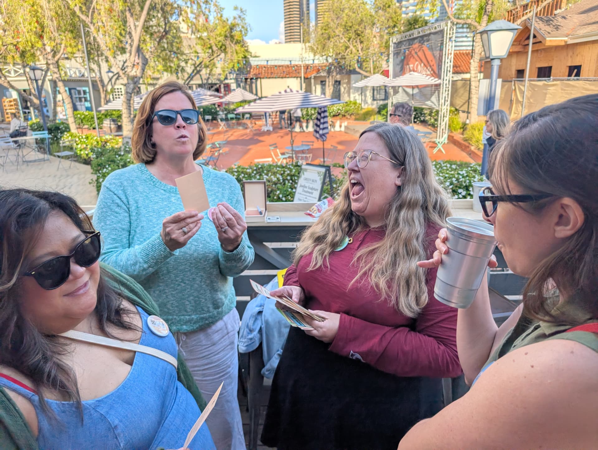 Four players laughing outdoors while holding cards in a sunny plaza.