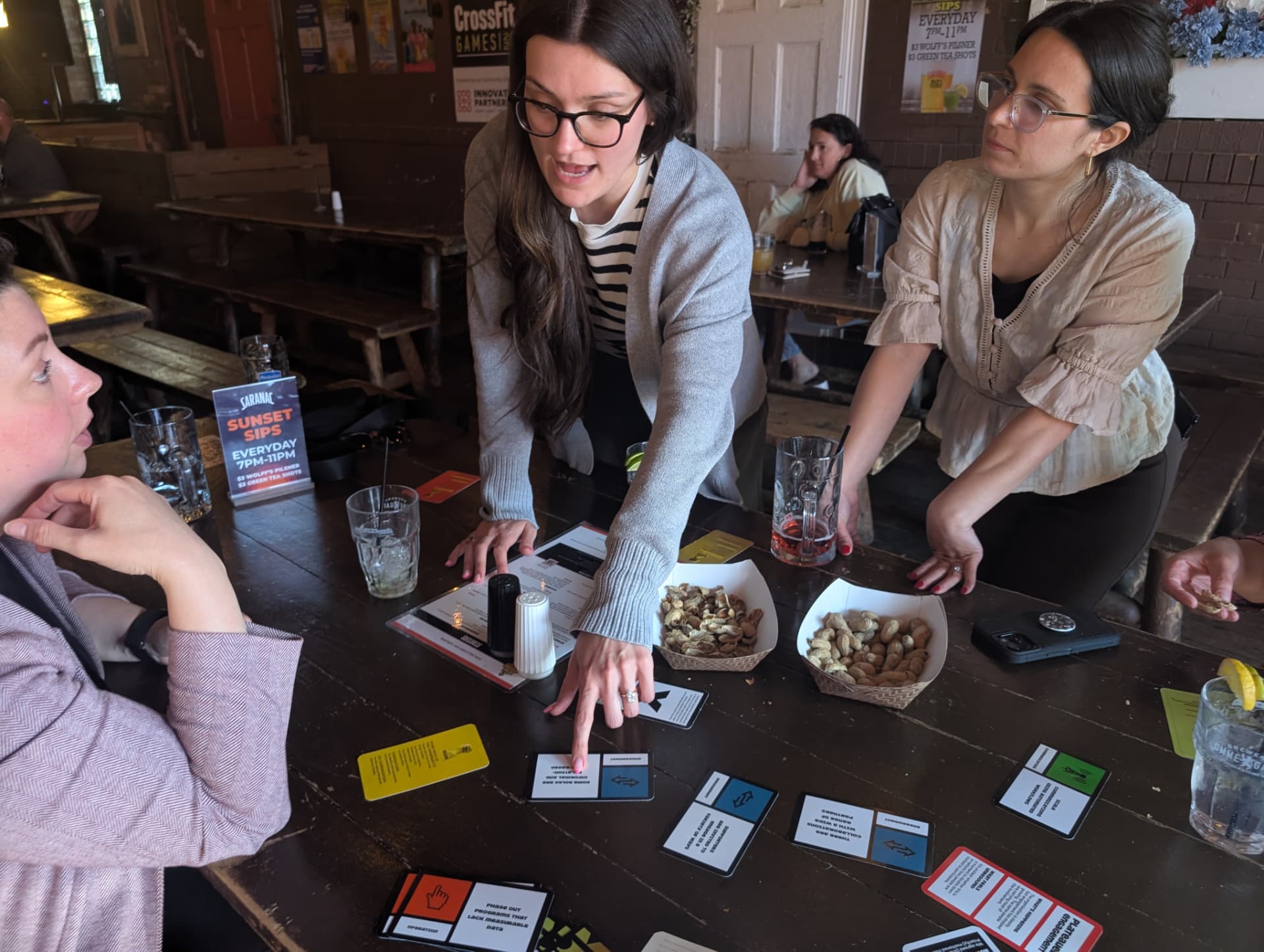 Three players lean over a long wooden tavern table pointing at cards in play.