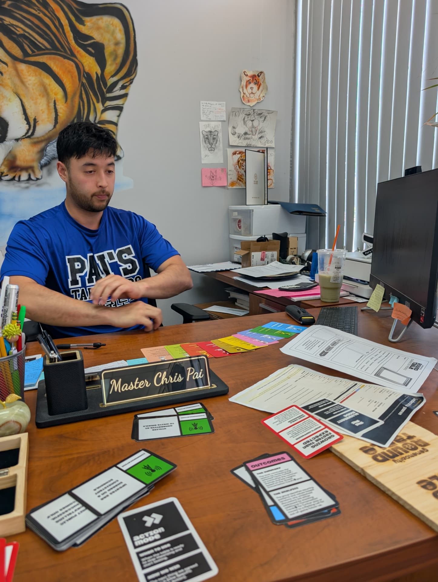 A player sits at an office desk with cards, character sheets and the wooden Roundtable box laid out.