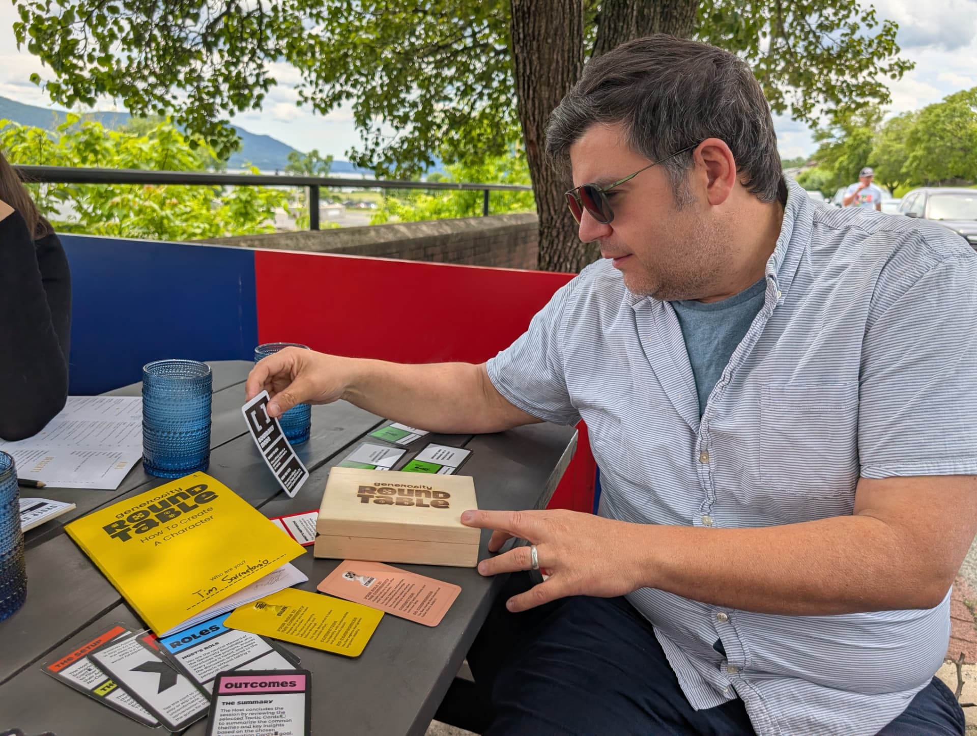 A player in sunglasses draws a card from the wooden Generosity Roundtable box at an outdoor table.