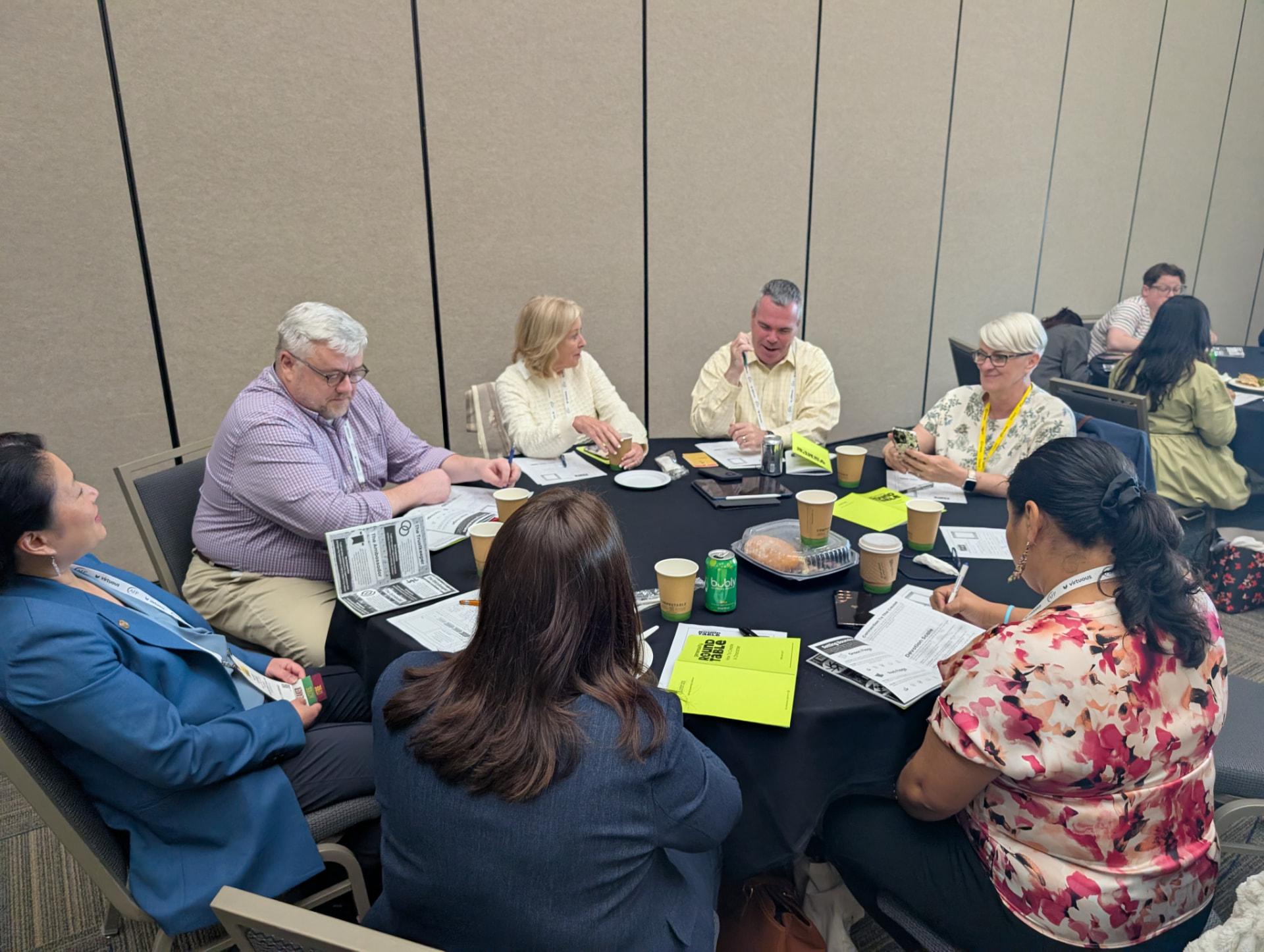 Eight players seated around a round conference table with character sheets and quick guides.