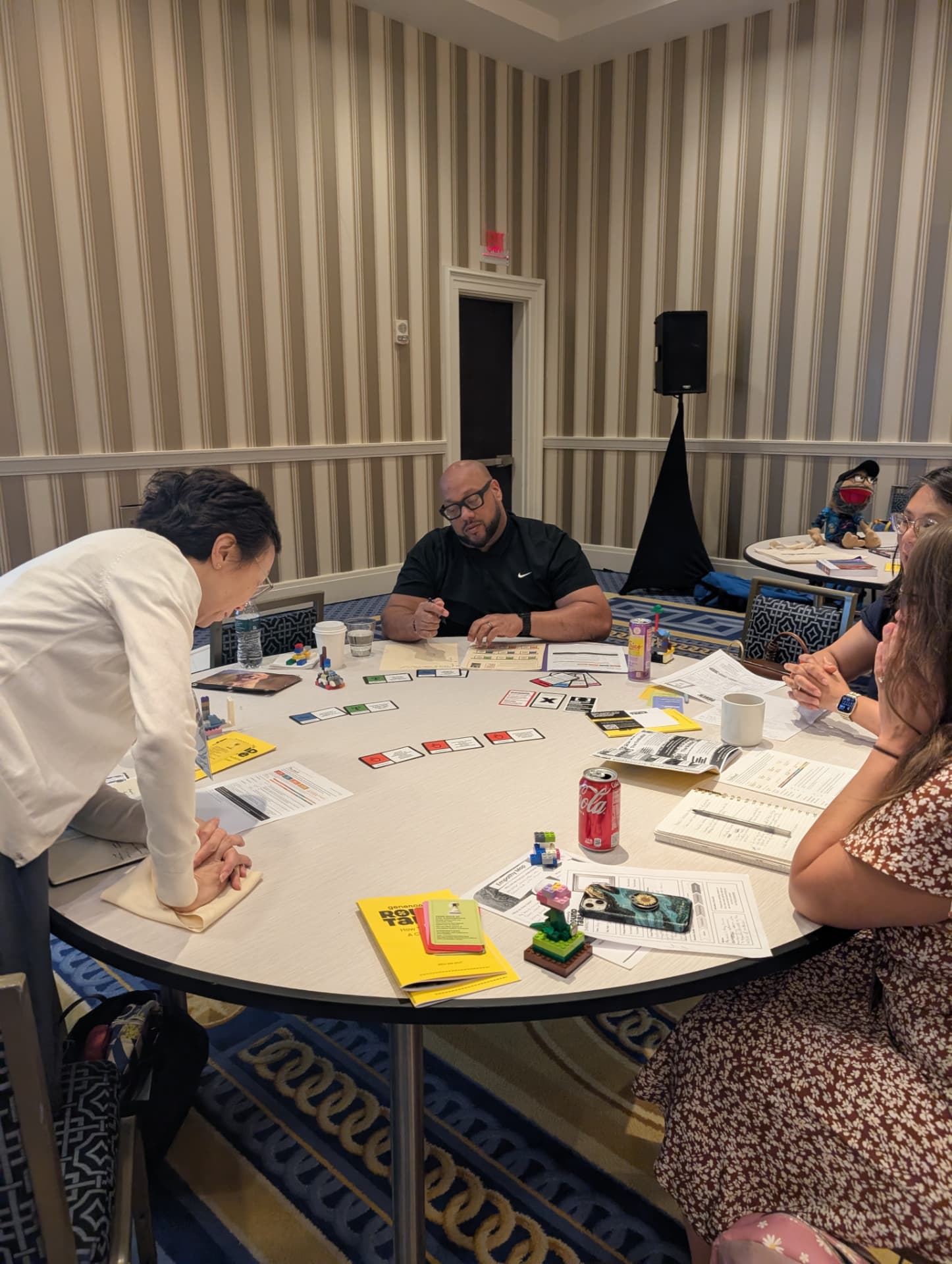 A Host leans over a ballroom table covered in cards, bricks and character sheets while three players consider a move.